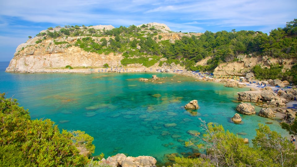 A coastal landscape with clear turquoise waters, rocky outcrops, and a hill covered in greenery.