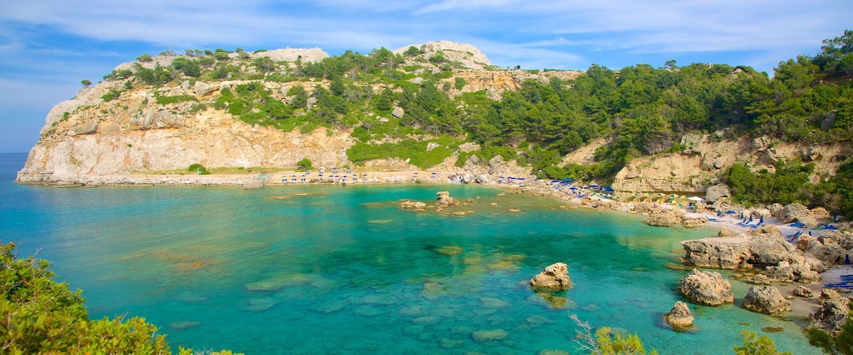 A coastal landscape with clear turquoise waters, rocky outcrops, and a hill covered in greenery.