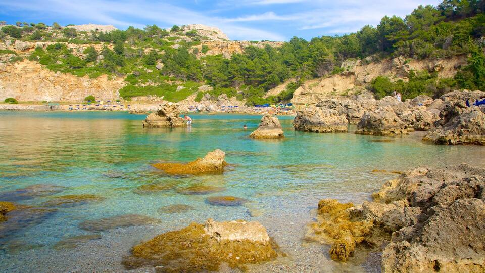 A rocky shoreline with clear turquoise water, surrounded by green hills and trees.