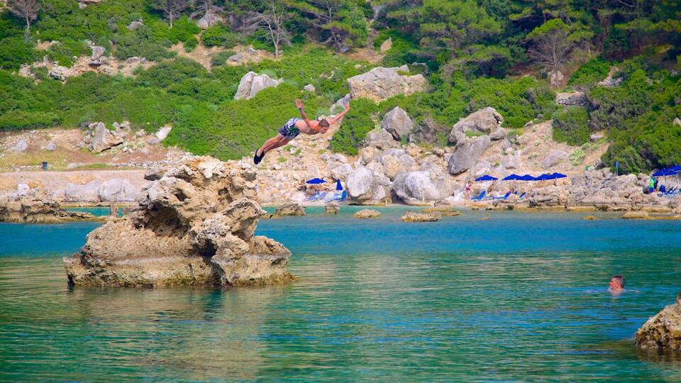 A man jumping off a rocky cliff into turquoise water, with a beach and umbrellas in the background.
