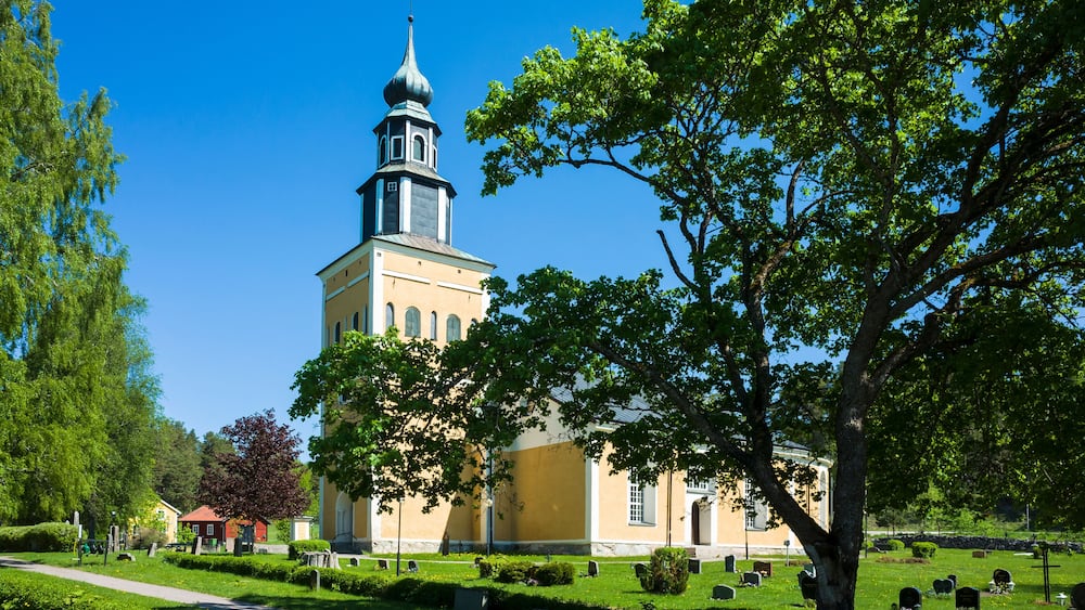 Yellow and white church building surrounded by a graveyard green lawn with gravestones under a large spreading tree under a blue sky, RamnÀs church in Sweden