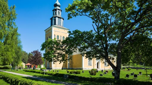 Yellow and white church building surrounded by a graveyard green lawn with gravestones under a large spreading tree under a blue sky, Ramnäs church in Sweden