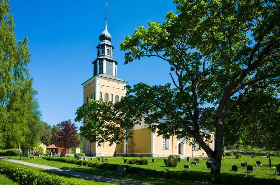 Yellow and white church building surrounded by a graveyard green lawn with gravestones under a large spreading tree under a blue sky, RamnÀs church in Sweden