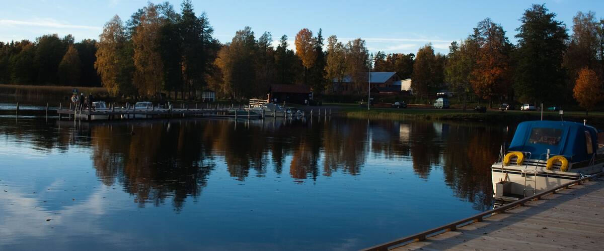 A calm lake with a dock, boats, and autumn trees in the background.