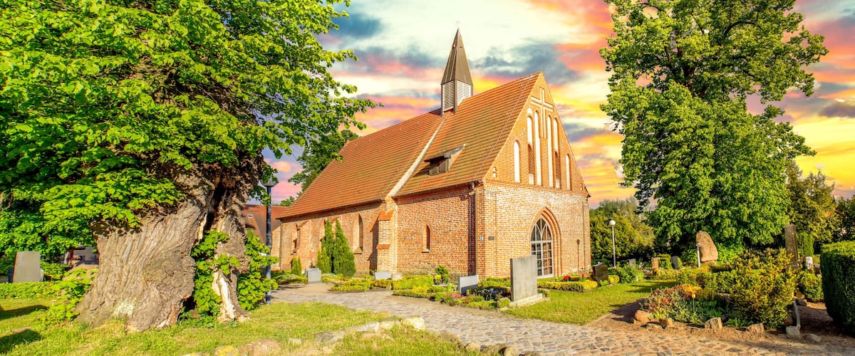 A brick church with a tall steeple, surrounded by lush greenery and a cobblestone path.