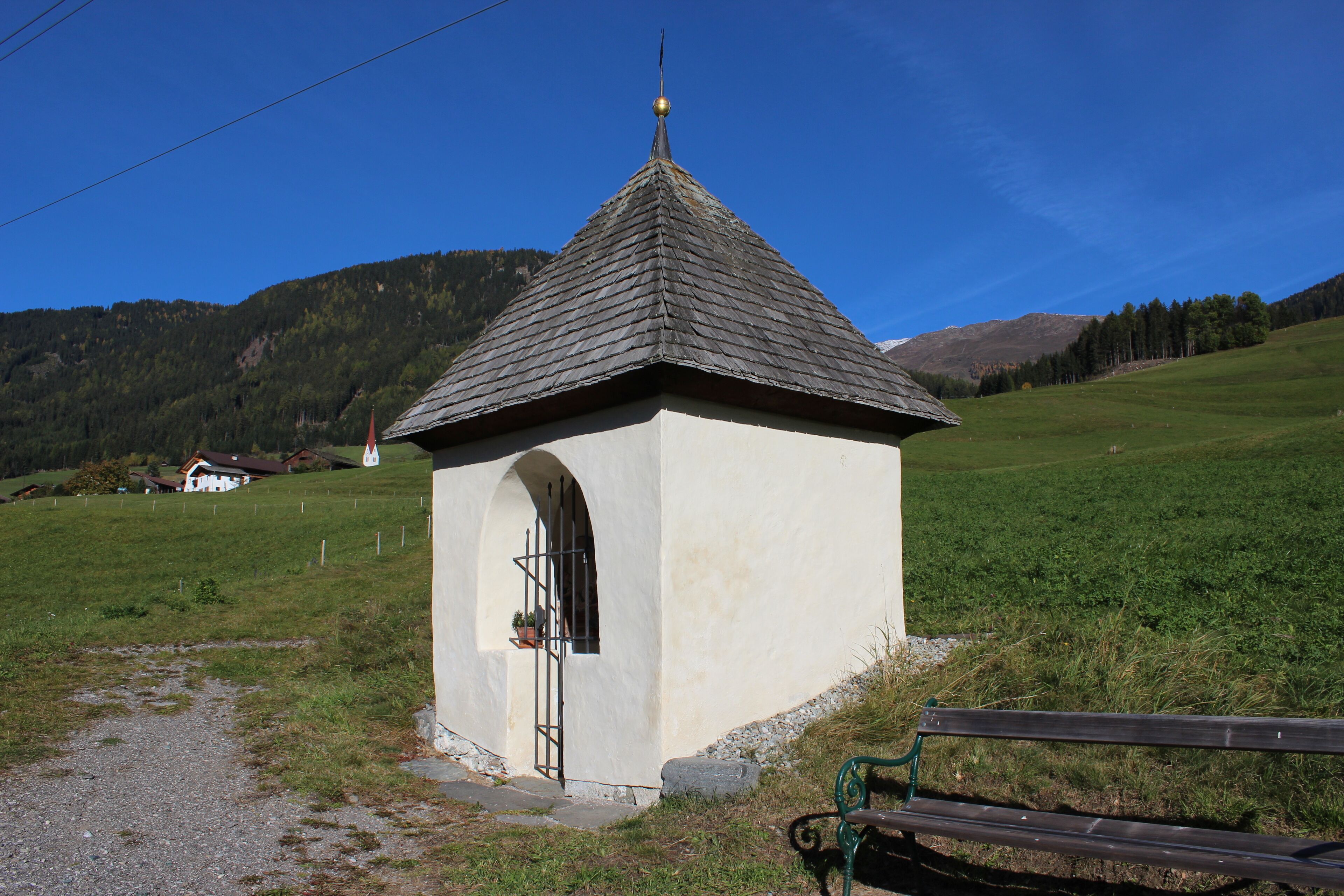 A small white chapel with a pointed roof and a bell tower, surrounded by a grassy field and mountains in the background.