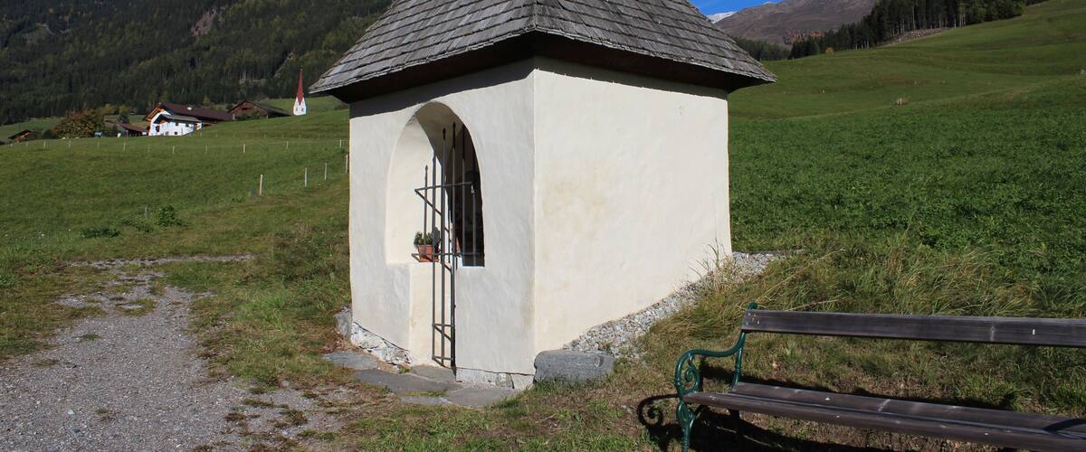 A small white chapel with a pointed roof and a bell tower, surrounded by a grassy field and mountains in the background.