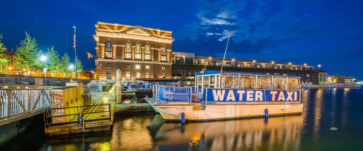 Un paseo marítimo con una embarcación de 'WATER TAXI' amarrada a un muelle, un edificio grande de arquitectura clásica y un río tranquilo que refleja las luces.