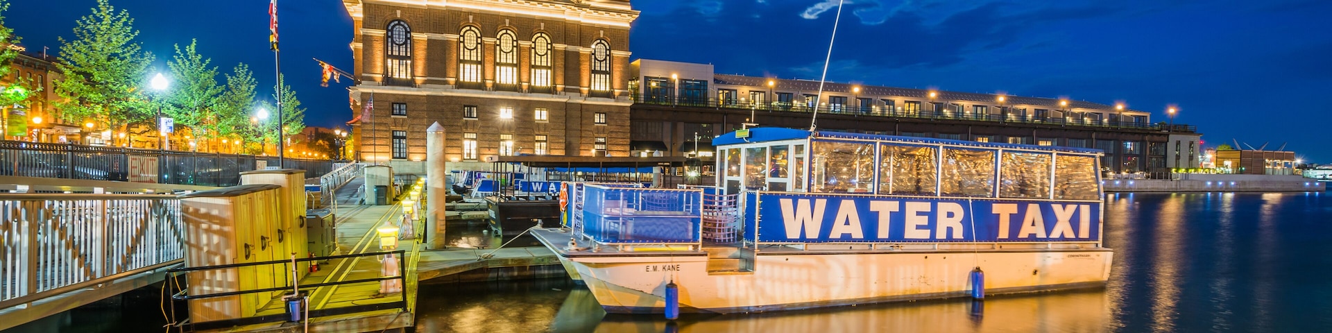 Ein Ufer mit einem 'WATER TAXI' Boot, das an einem Steg festgemacht ist, ein großes Gebäude mit klassischer Architektur und ein ruhiger Fluss, der die Lichter spiegelt.
