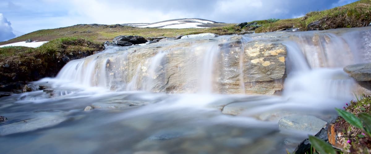 A waterfall cascading over rocks with a snow-capped hill in the background.