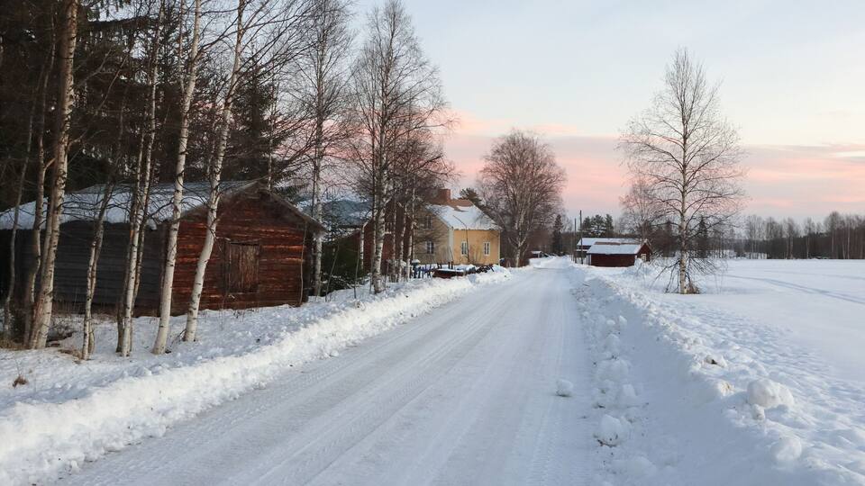 A snow-covered rural road flanked by leafless trees and wooden buildings.