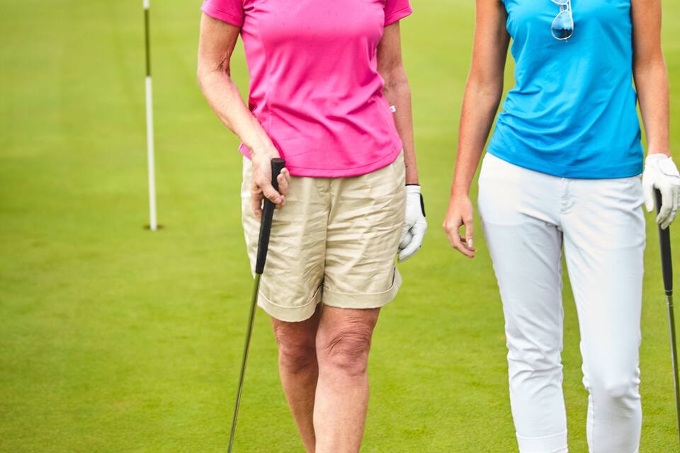 Twee personen op een golfbaan, met een vlag, water en huizen op de achtergrond.