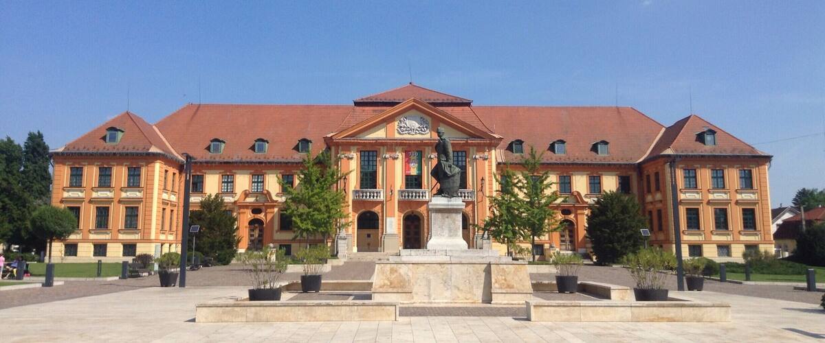 A large, historic building with a red roof and multiple windows. In front, there is a statue on a pedestal and several potted plants.