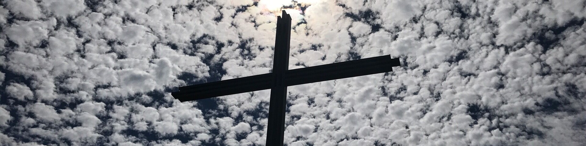 Uma cruz silhuetada contra um céu repleto de nuvens, com o sol a brilhar.