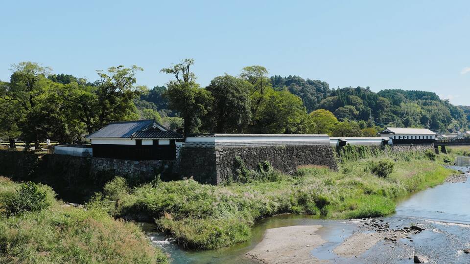 A riverbank with a traditional building, a stone wall, and a forested hill in the background.
