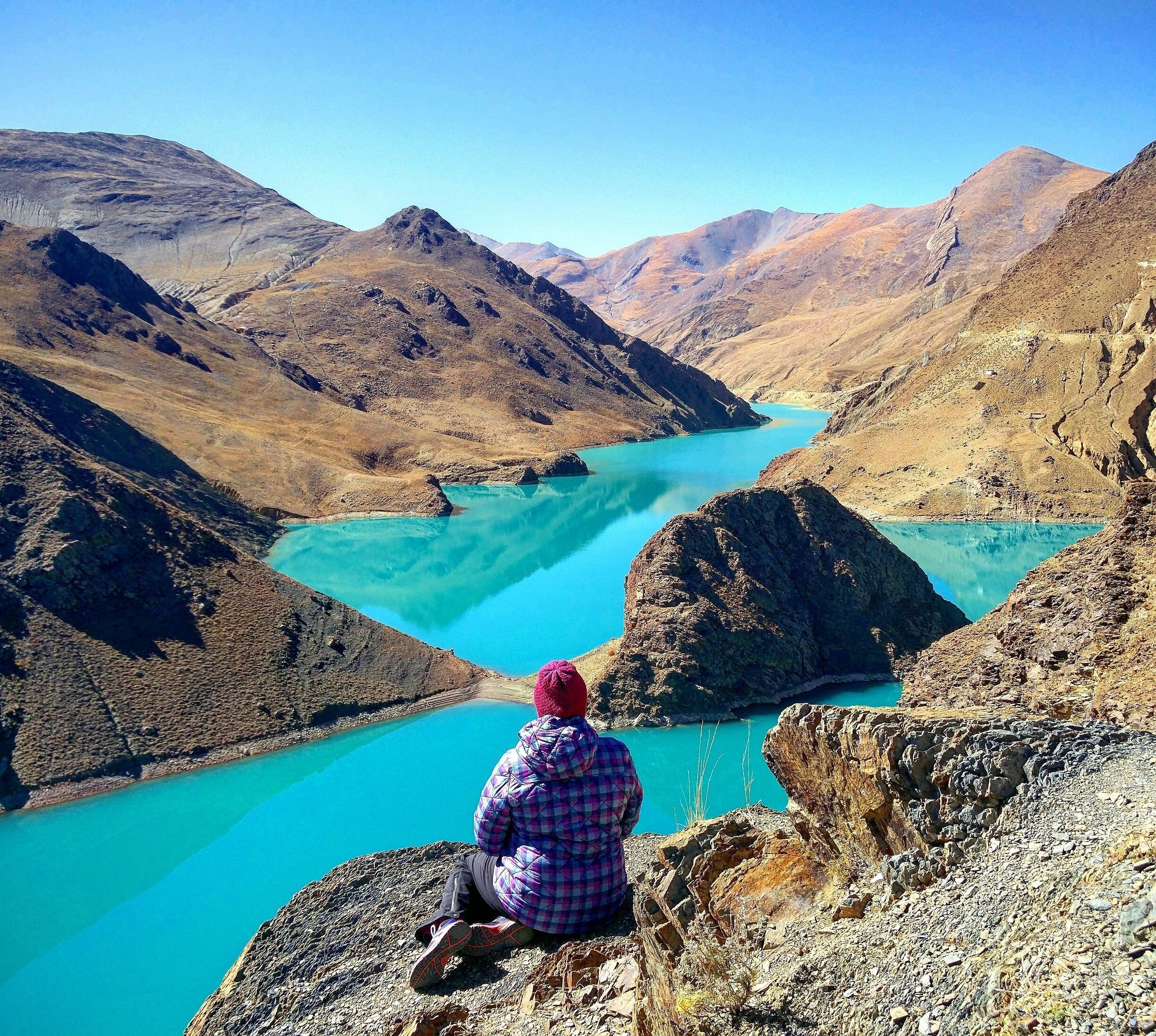A person sitting on a rocky outcrop overlooking a turquoise lake surrounded by mountains.