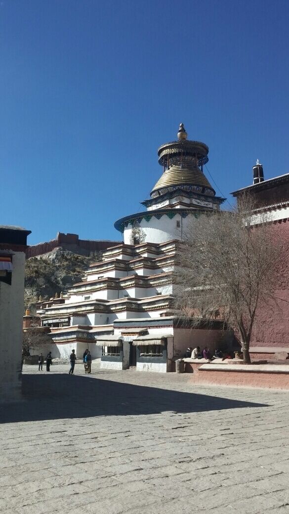 A traditional Tibetan stupa with multiple tiers and a golden top, surrounded by a courtyard with people and a tree.