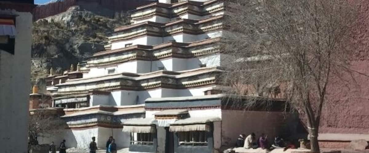 A traditional Tibetan stupa with multiple tiers and a golden top, surrounded by a courtyard with people and a tree.