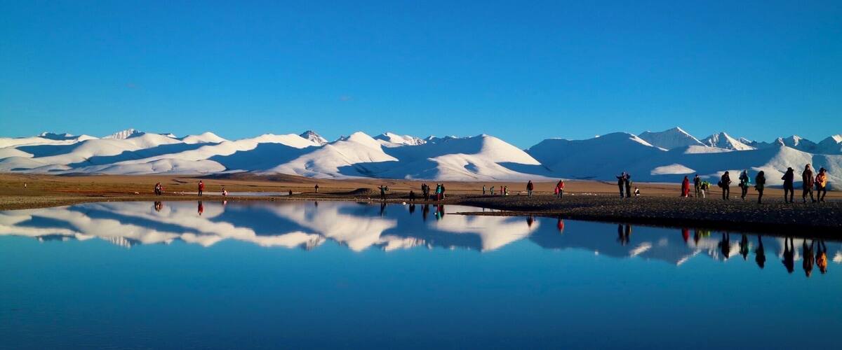 Un grupo de personas de pie junto a un lago que refleja, con montañas nevadas al fondo.