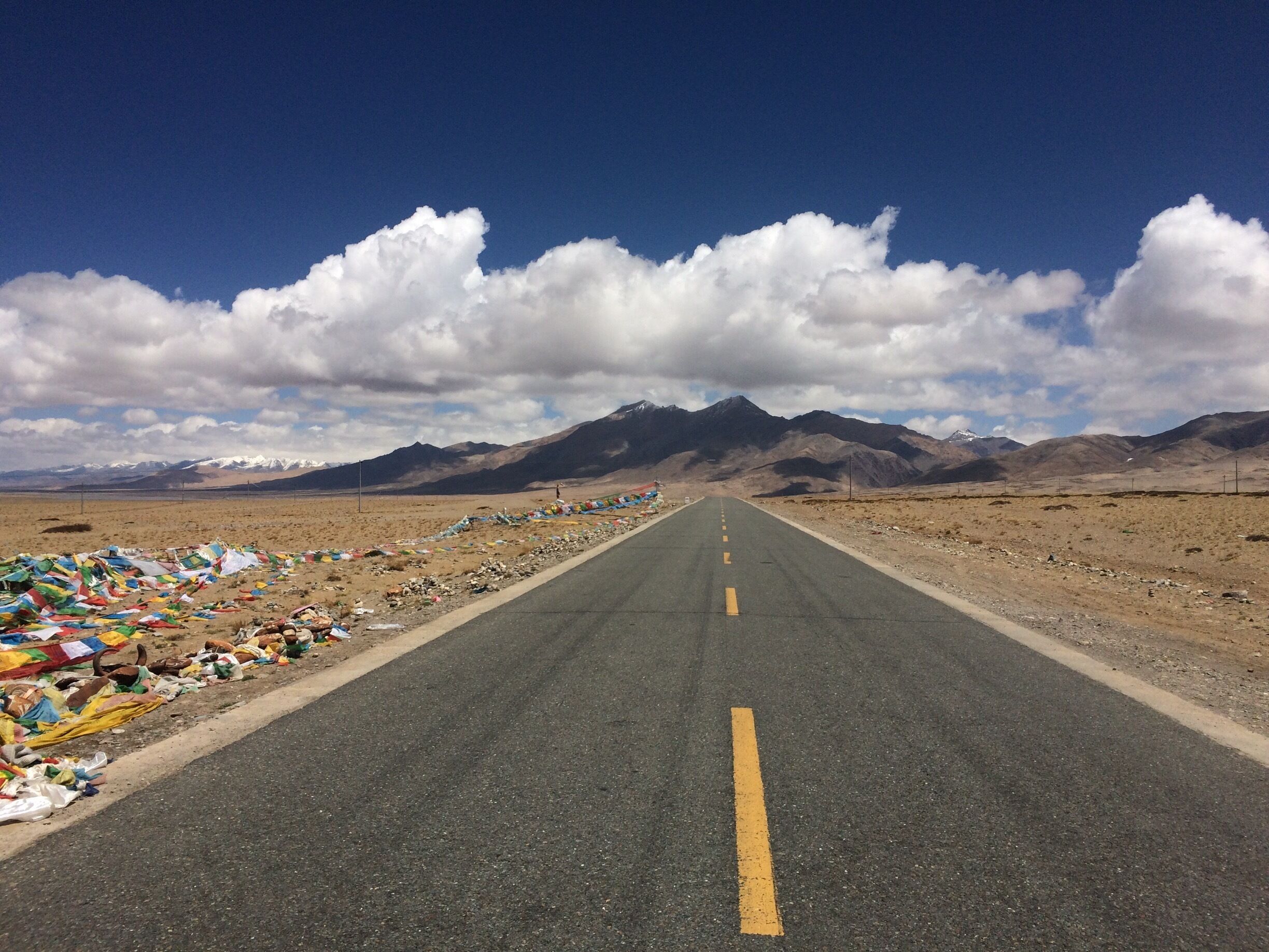 A road with colorful prayer flags on the left, leading to a mountain range under a clear sky.