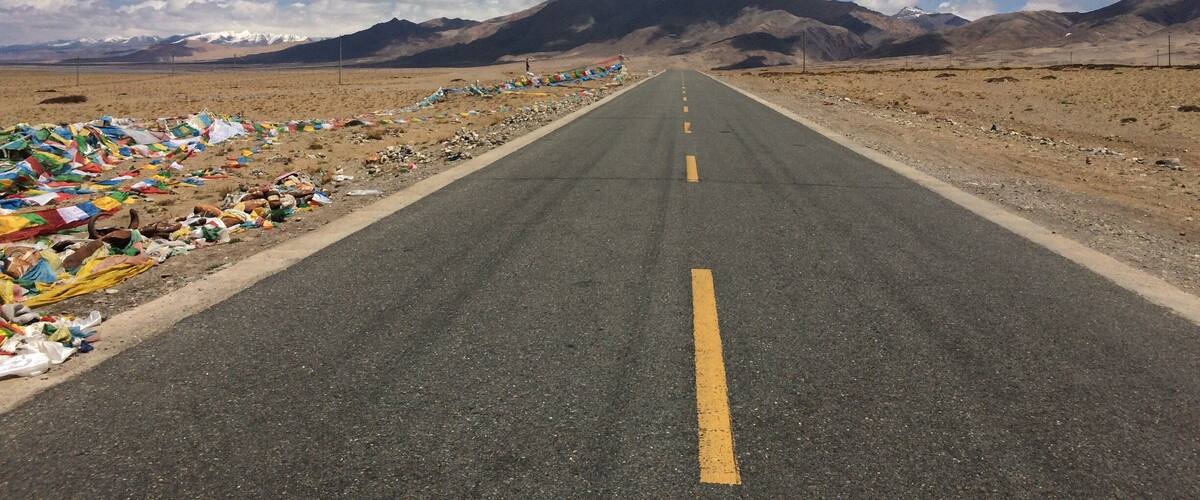 A road with colorful prayer flags on the left, leading to a mountain range under a clear sky.