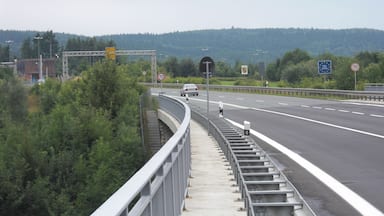 A highway with a speed limit sign of 70 km/h, a pedestrian bridge, and a forested hillside in the background.