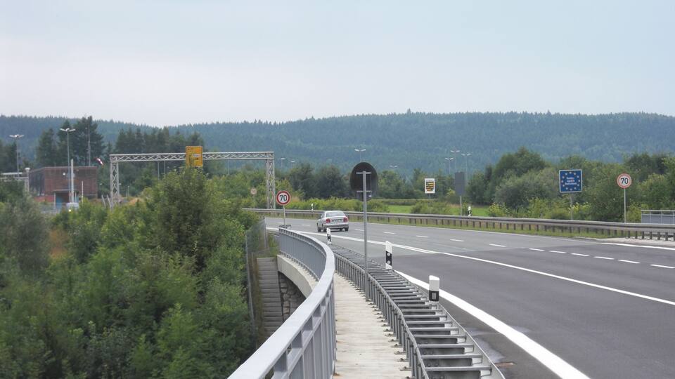 A highway with a speed limit sign of 70 km/h, a pedestrian bridge, and a forested hillside in the background.
