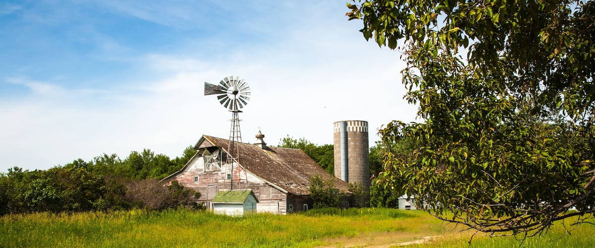 A rustic barn with a windmill, a silo, and a dirt path in a lush green field.