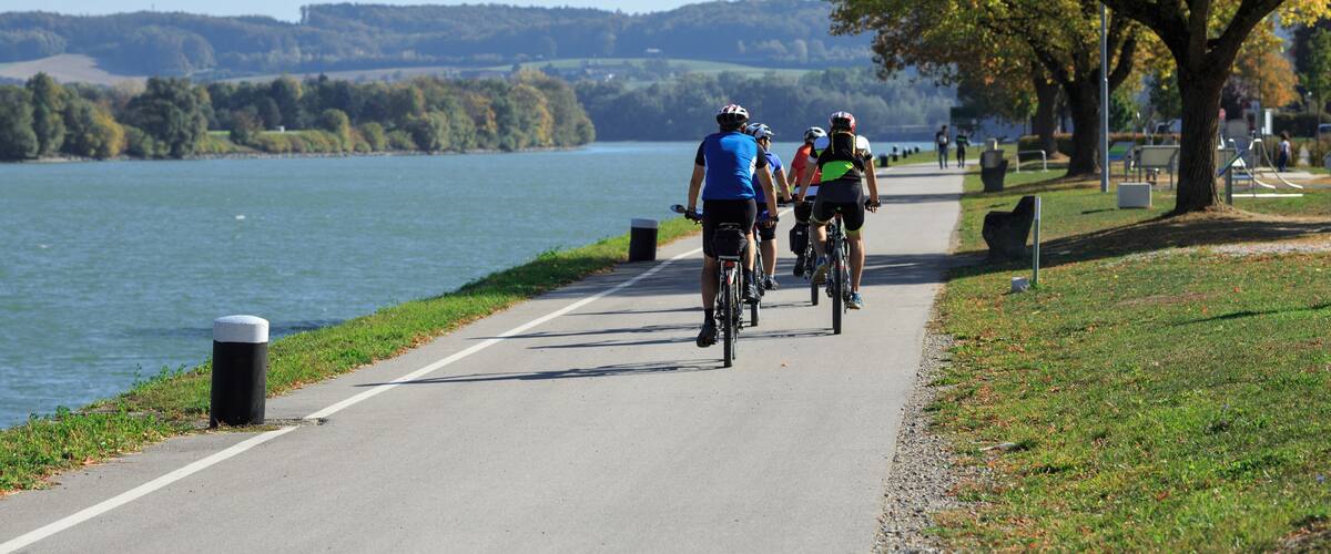 A group of cyclists on a paved path by a lake with trees and hills in the background.