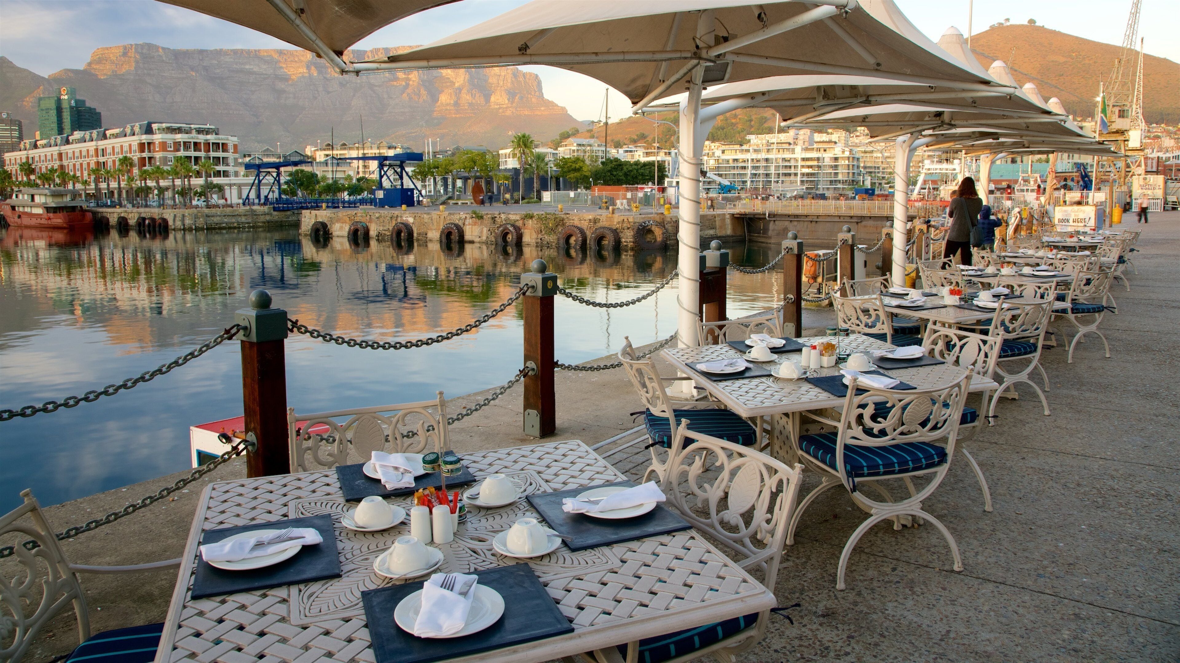 A waterfront dining area with tables and chairs, a chain barrier, and a view of a cityscape with buildings and mountains in the background.
