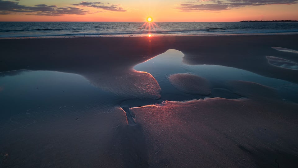A sunset over a beach with a reflective water surface and a silhouette of a bridge.