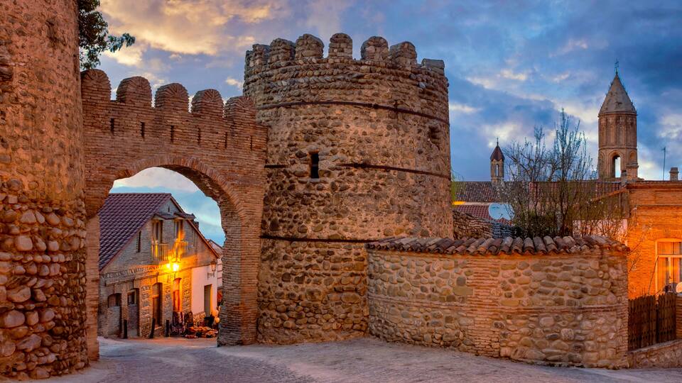A stone castle gate, a church with a bell tower, and a cobblestone street.