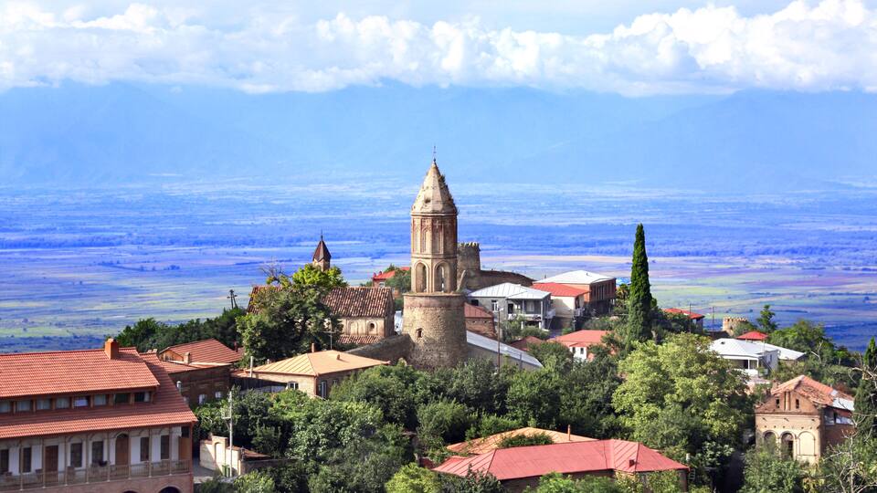 A historic town with a prominent church tower, lush greenery, and a mountainous backdrop.