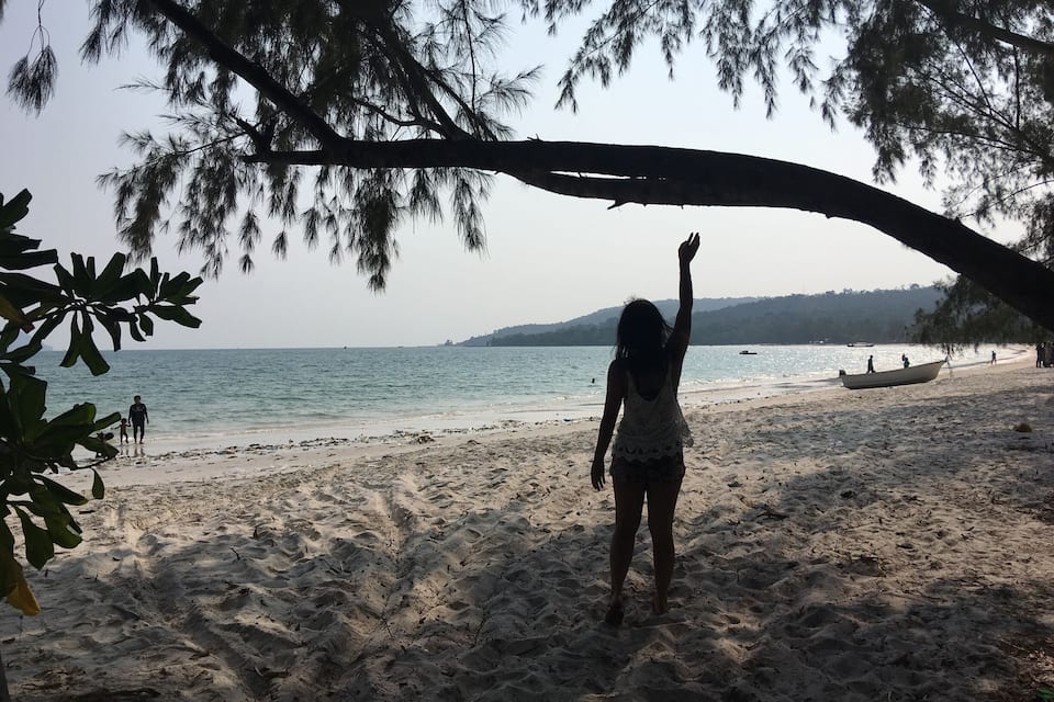 A beach with a boat on the sand, a person raising their hand, and trees in the background.