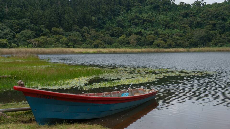 A small blue boat with red trim, labeled "ZAPASSA," docked by a calm lake with dense forest in the background.
