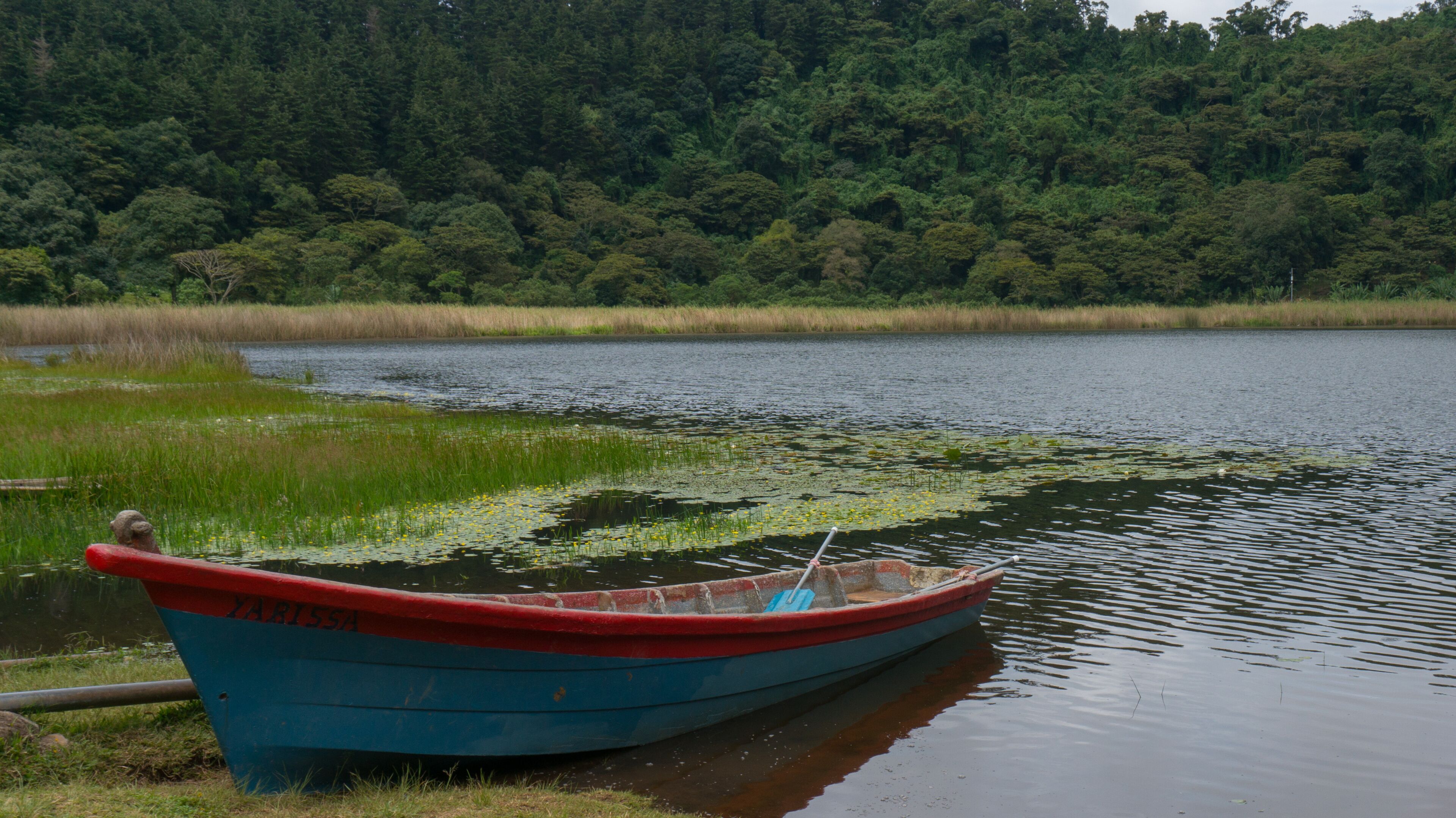A small blue boat with red trim, labeled "ZAPASSA," docked by a calm lake with dense forest in the background.