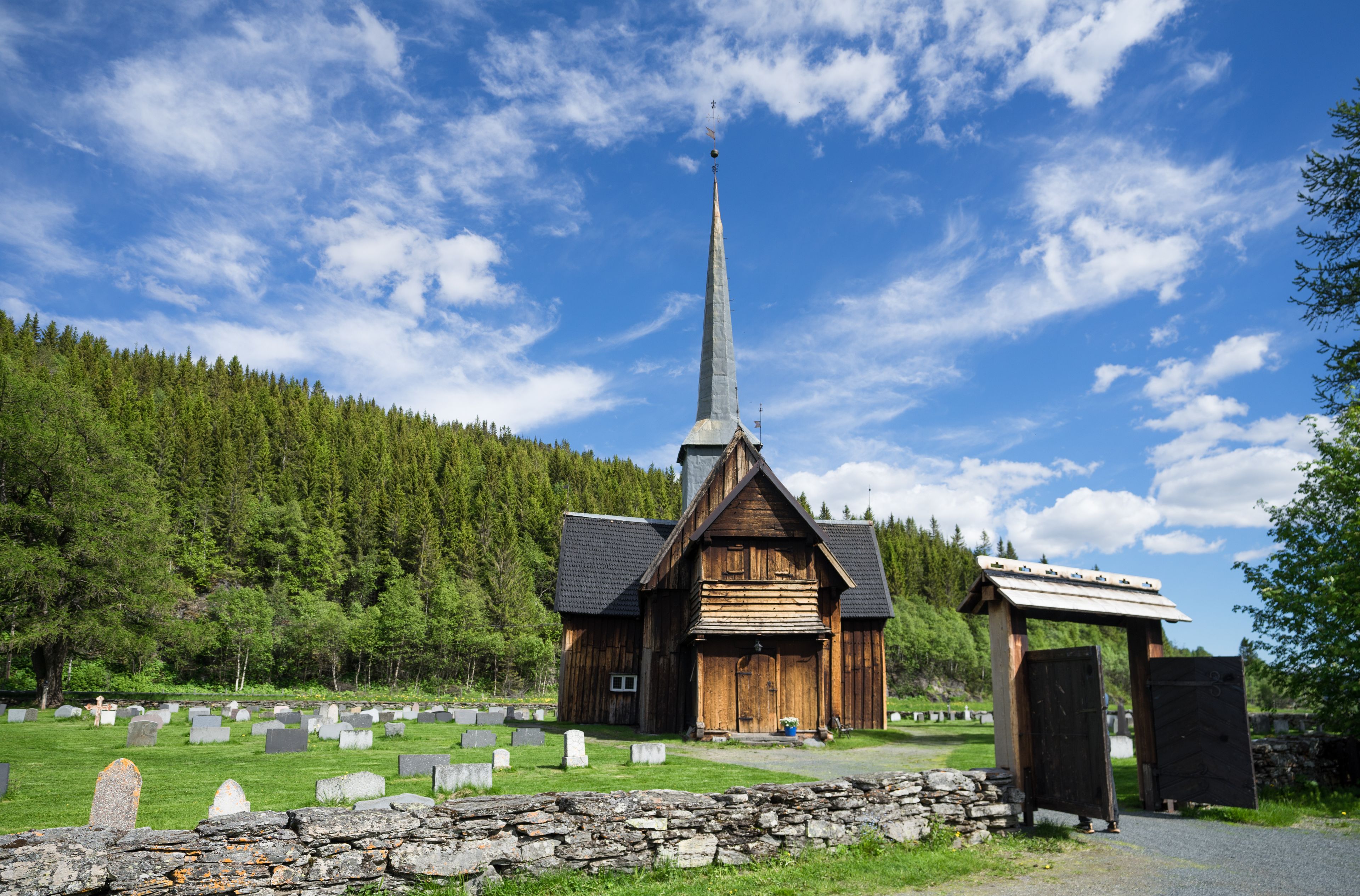 Une église en bois, dotée d’un clocher imposant, entourée d’un cimetière où se trouvent des pierres tombales et un mur de pierre.