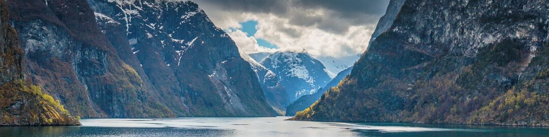 Un lago tranquilo rodeado de montañas agrestes con picos nevados bajo un cielo nublado.