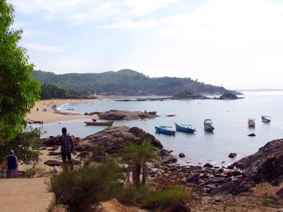 A rocky shoreline with boats, a sandy beach, and a forested hill in the background.