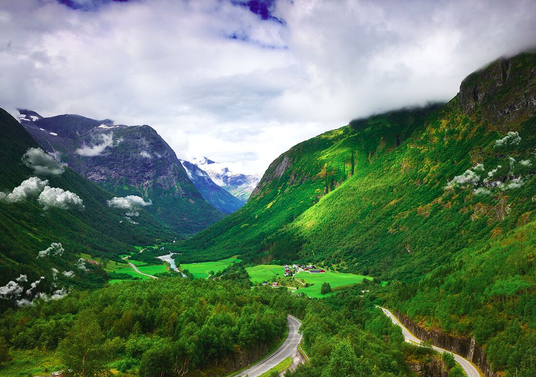 A valley with a winding road, green fields, and mountains in the background.