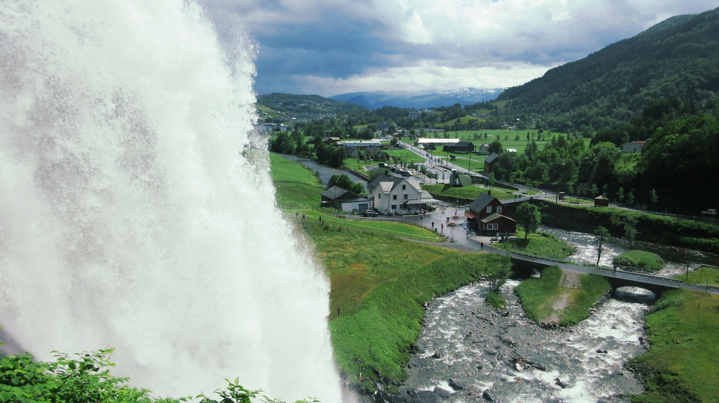 A waterfall cascading down a rocky cliff, a river flowing through a valley, and a small village with houses and a bridge.