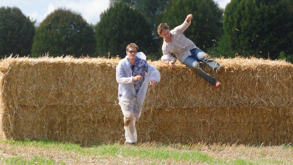 Two people interacting with a hay bale in a field with trees in the background.