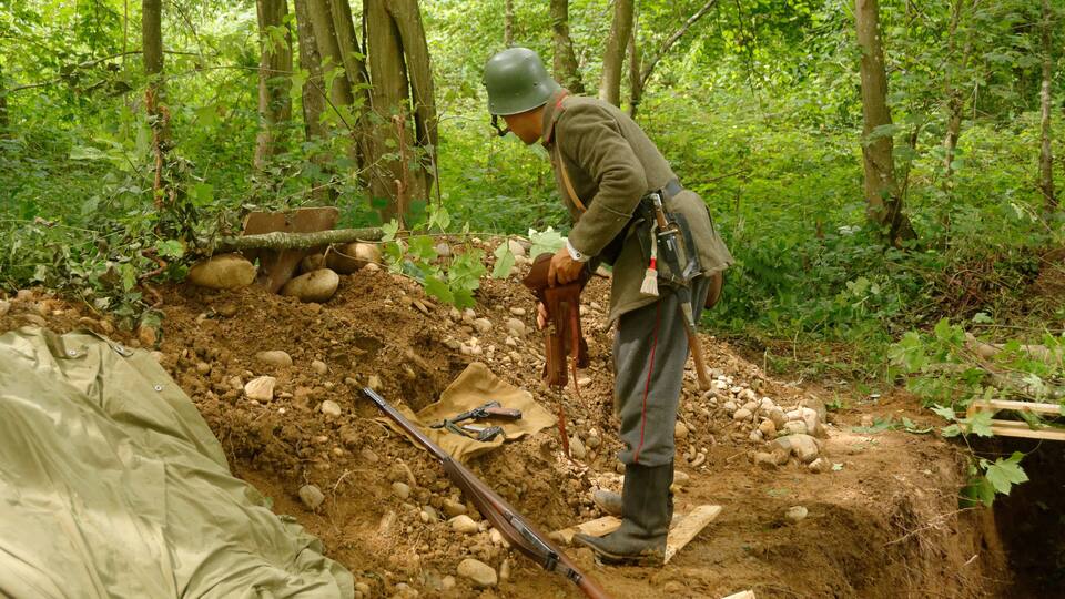 A soldier in a forest with a trench, a rifle, and a shovel.