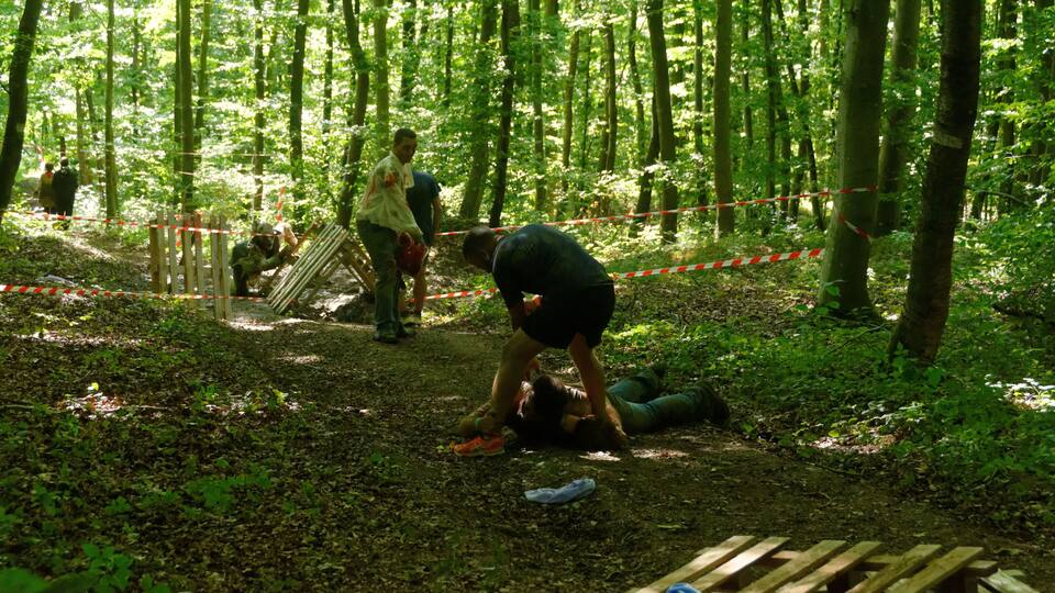 A forest with a person lying on the ground and another person attending to them.
