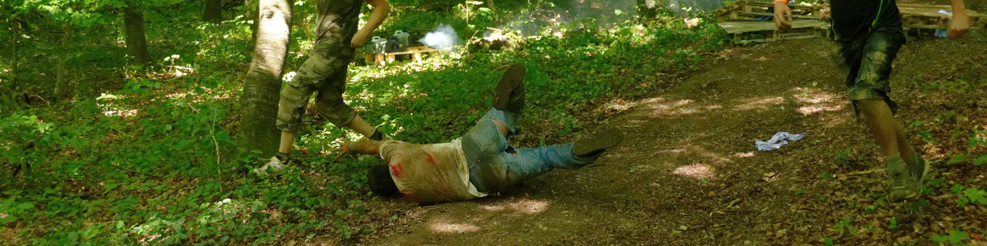 A forested area with a dirt path, a person lying on the ground, and another person running.