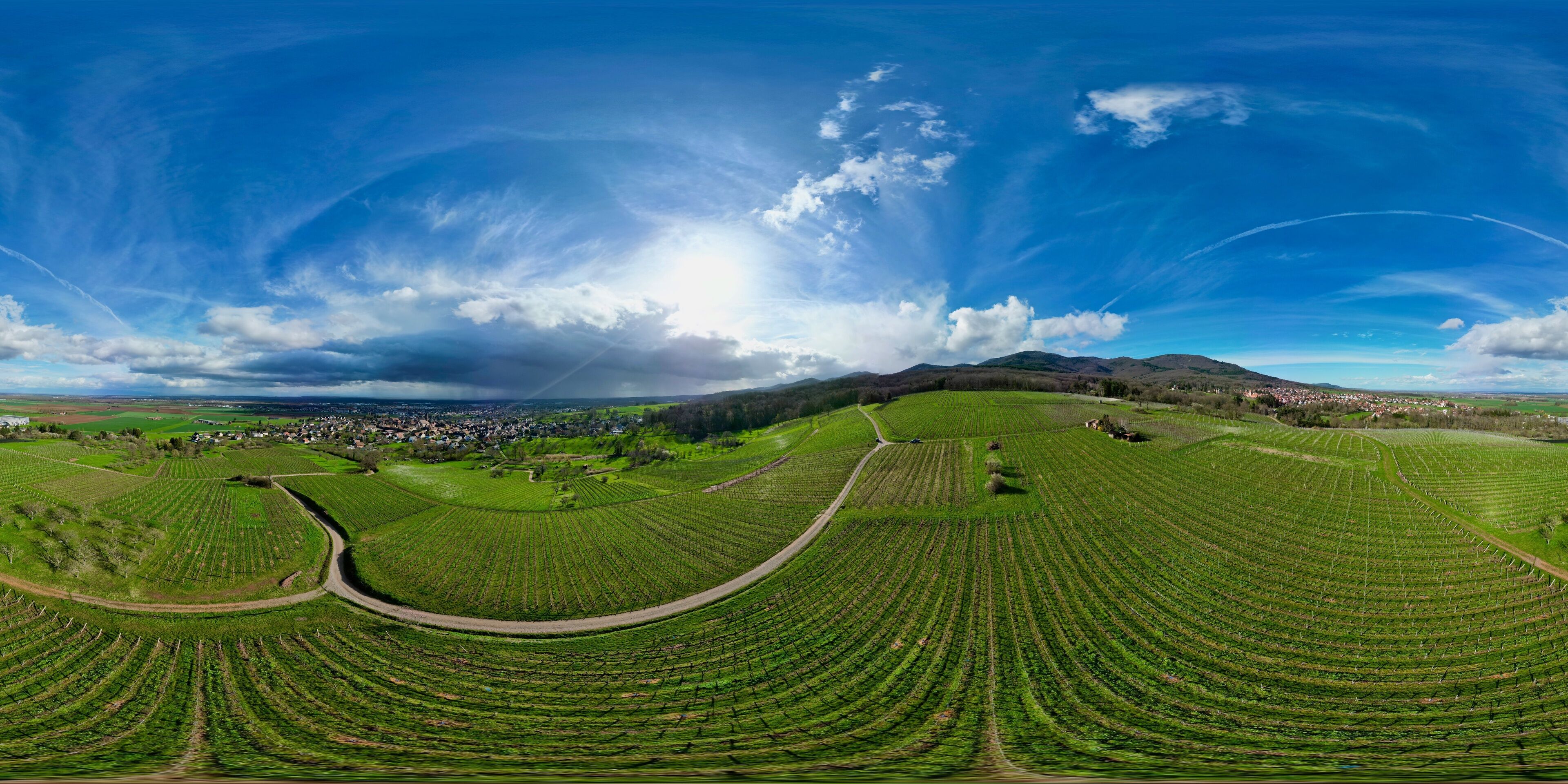 360 panorama aerial drone view above the Uffholtz and Wattwiller villages green vineyard hills in a Alsatian valley by sunny day with blue sky, dark stormy clouds in the distance, with paths