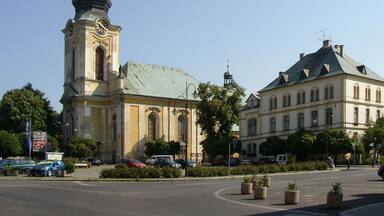 A church with a tall steeple and a clock, a roundabout with potted plants, and a large building with a green roof.