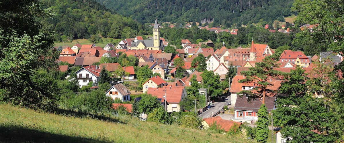 A village nestled in a valley with red-roofed houses, surrounded by lush green hills and a prominent church tower.