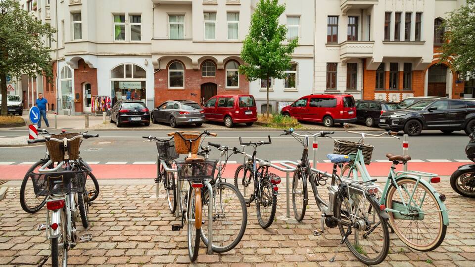Eine Reihe von Fahrrädern steht auf einer Kopfsteinpflasterstraße, im Hintergrund sind Autos und ein roter Transporter zu sehen.