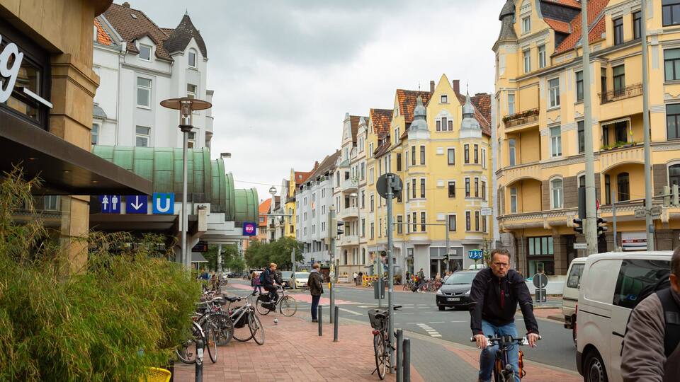 Eine Straße mit Fahrrädern, Fußgängern und Autos. Es gibt Gebäude mit bunten Fassaden und eine Bushaltestelle.